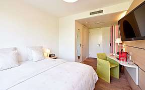 Hell eingerichtetes Hotelzimmer mit Doppelbett, grünem Stuhl und Wandfernseher