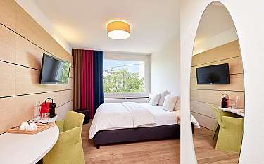 Helles Hotelzimmer mit Doppelbett, grünem Stuhl, Fernseher und großem Spiegel