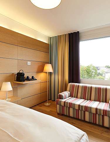 Helles Hotelzimmer mit Bett, Sofa und Fensterblick.